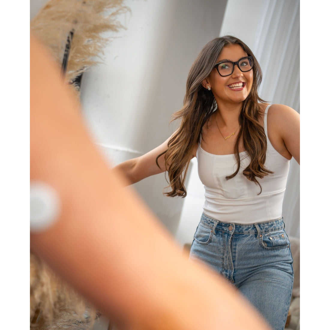 Woman wearing a white tank top and blue jeans, smiling in a room with a couch and decorative items.