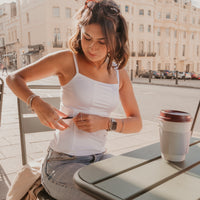 Woman sitting at an outdoor table with a coffee cup, surrounded by buildings and street wearing a white Mude Camisole.